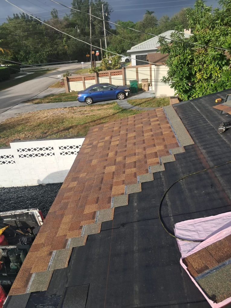 Roof being shingled with brown asphalt shingles; a blue car is parked in the street below.