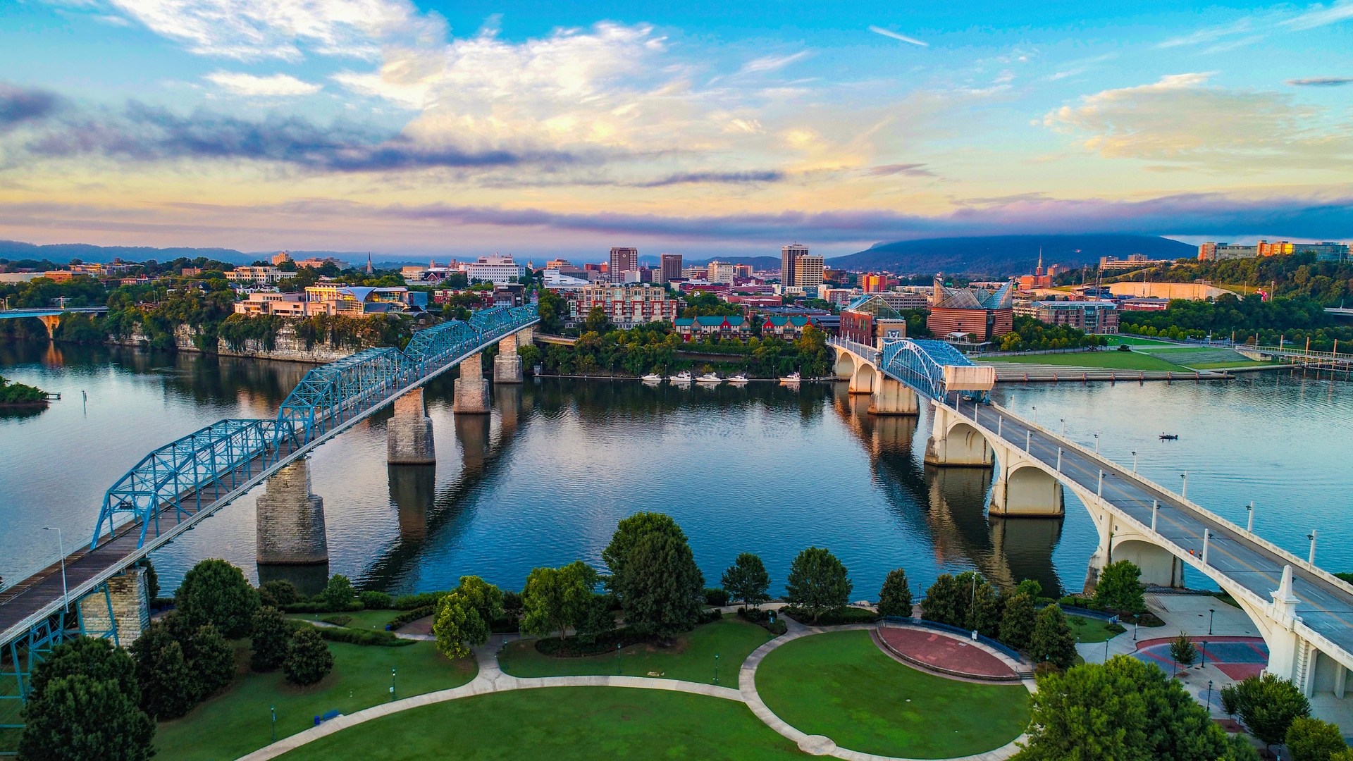 Aerial view of two bridges spanning a river at sunset, with a city skyline and park in the foreground.