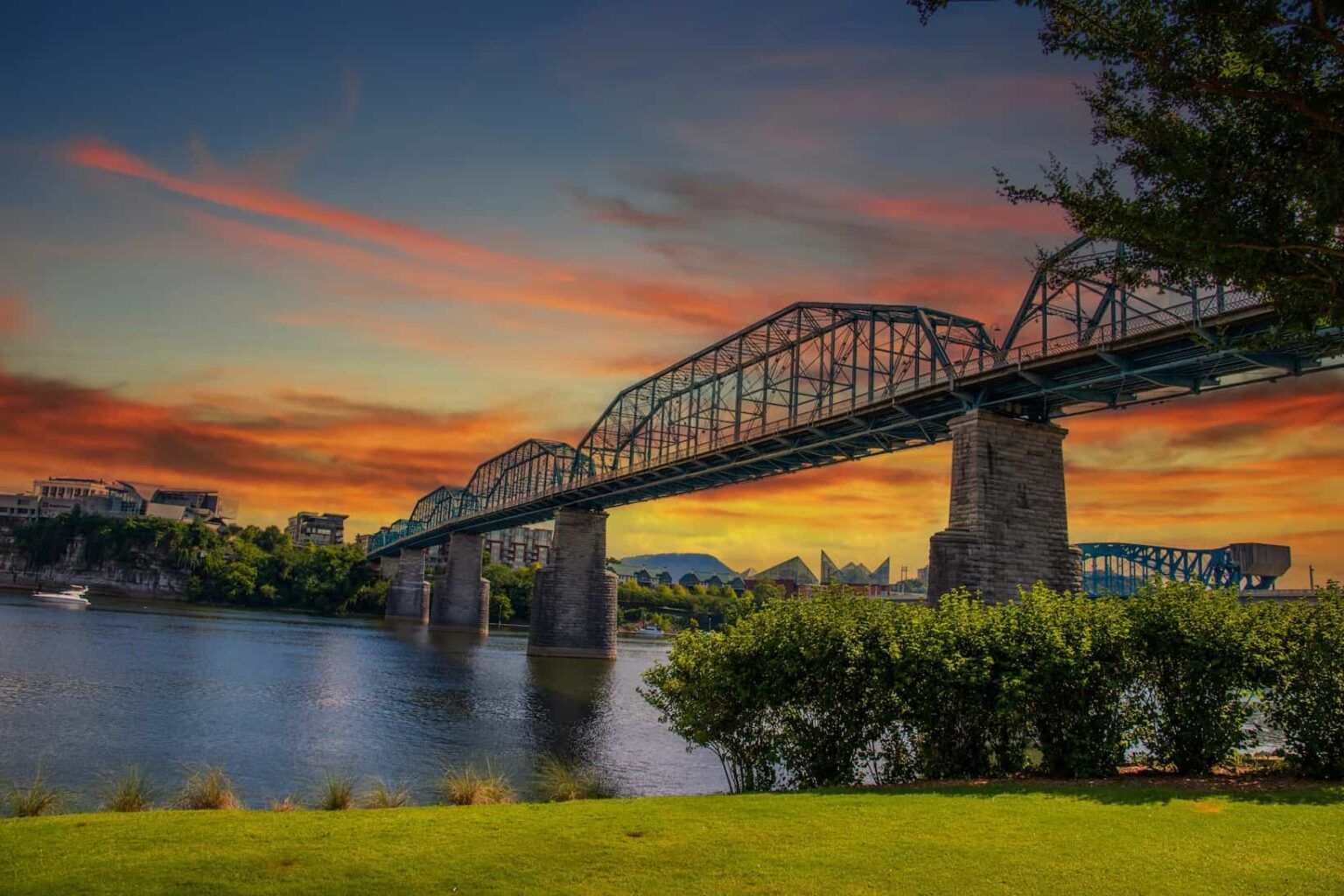 Sunset over a steel bridge spanning a river, with orange sky reflections and green grass in the foreground