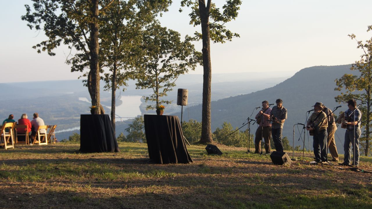 People standing on a grassy hill at sunset, with trees, chairs, and a distant mountain view.