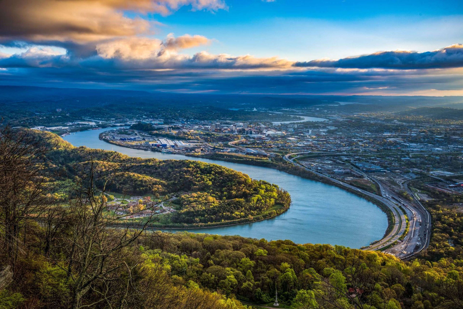 River winding through a valley at sunset, with hills, city lights, and dramatic clouds.