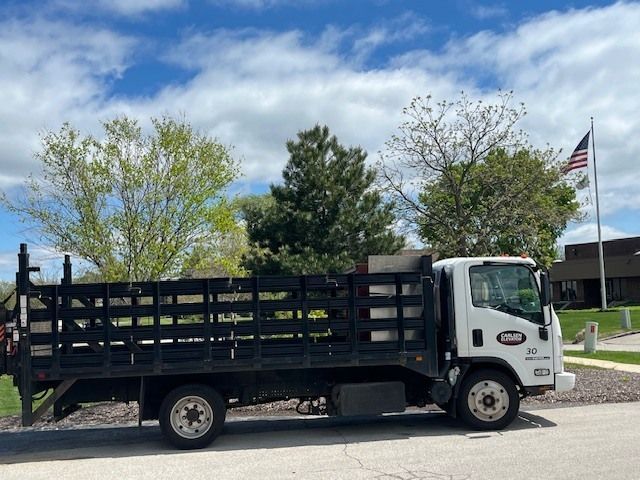 Black flatbed truck parked outdoors near trees and a flagpole under a cloudy sky