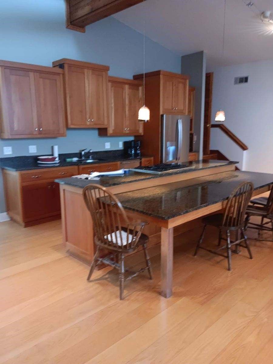 Open kitchen with wooden cabinets, island table, and chairs in a bright hardwood-floored room