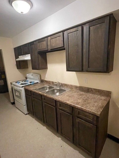 Small kitchen with dark wood cabinets, brown countertops, white stove, and stainless sink in an apartment.