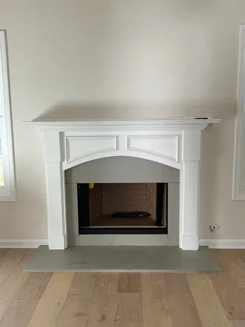 White decorative fireplace mantel with a dark firebox in a light-colored room, on a gray hearth.