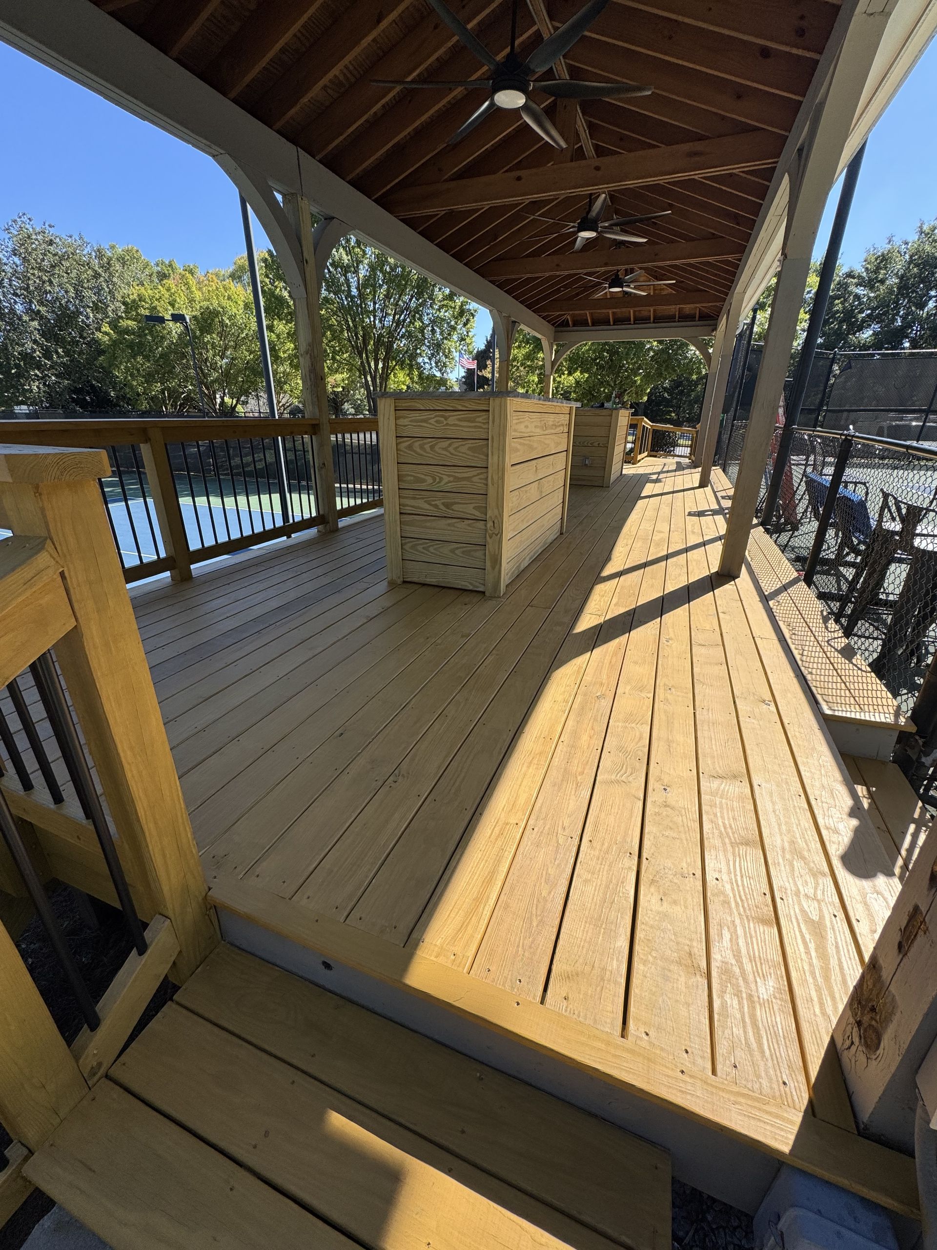 A sunlit wooden deck with a covered pavilion, wooden planter boxes, and railings overlooking a bright outdoor area.