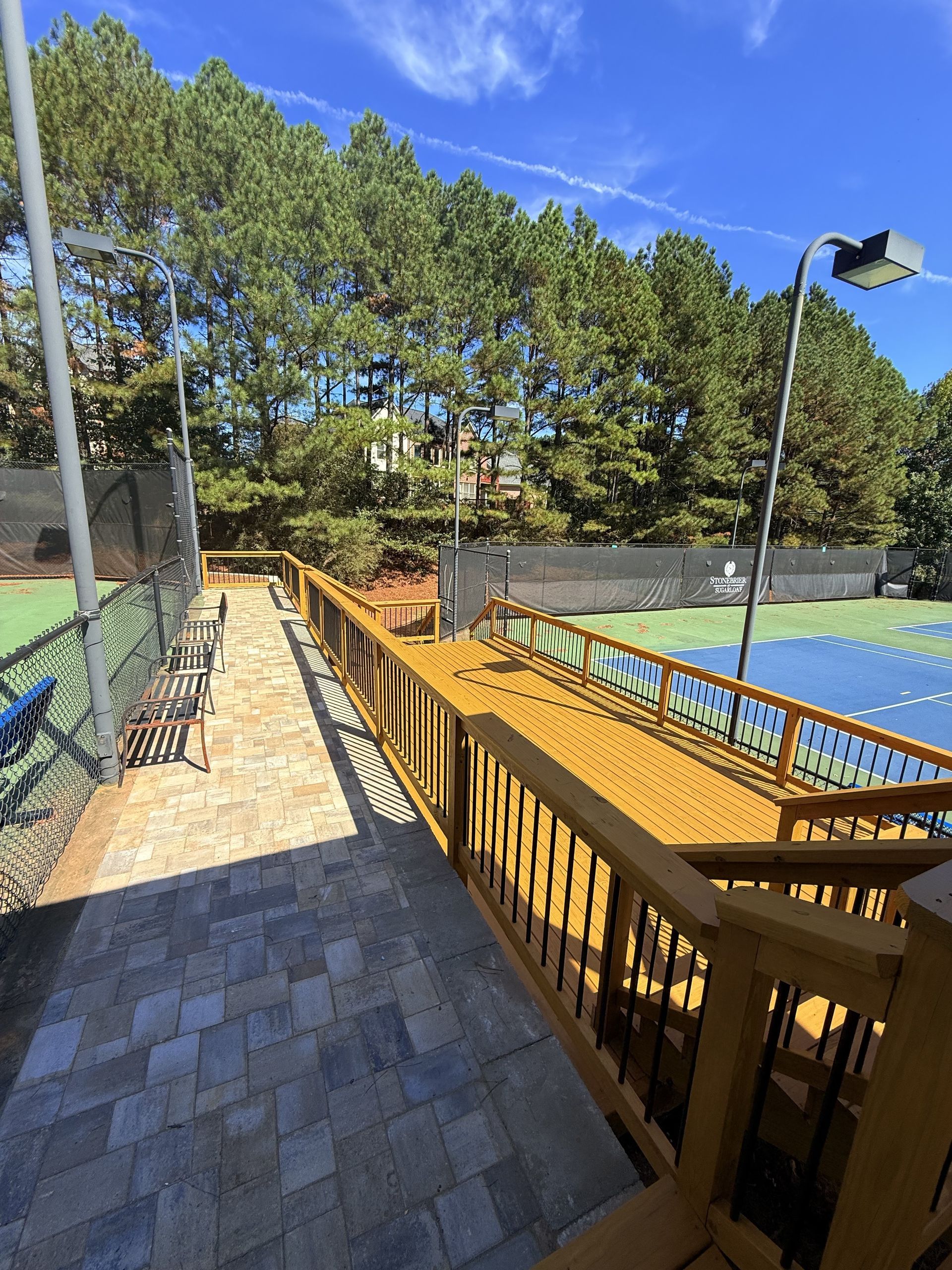 A wooden deck walkway with railings overlooking a blue and green tennis court on a sunny day with trees in the background.