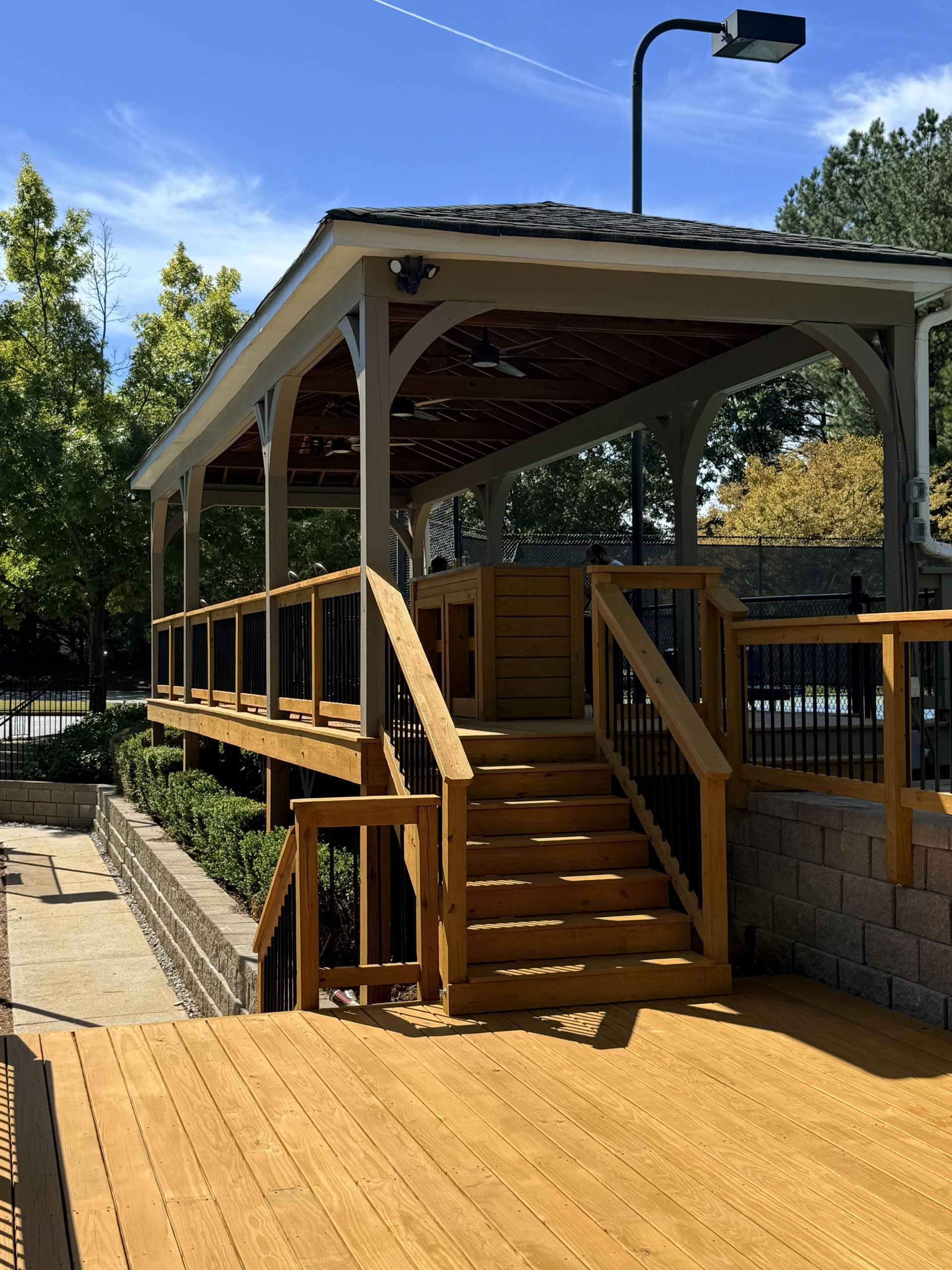 A raised wooden deck with a staircase leading up to a covered pavilion structure under a bright, sunny sky.