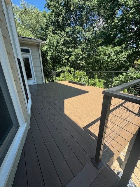 A modern elevated deck with dark brown composite boards, a metal cable railing, and a view of lush green trees.