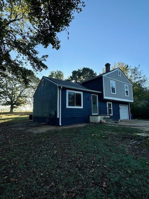 Blue two-story house with white trim in a grassy yard under trees, viewed from the side.