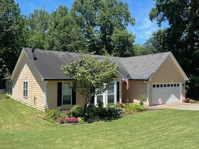 Single-story beige house with garage, front lawn, and trees under a bright blue sky