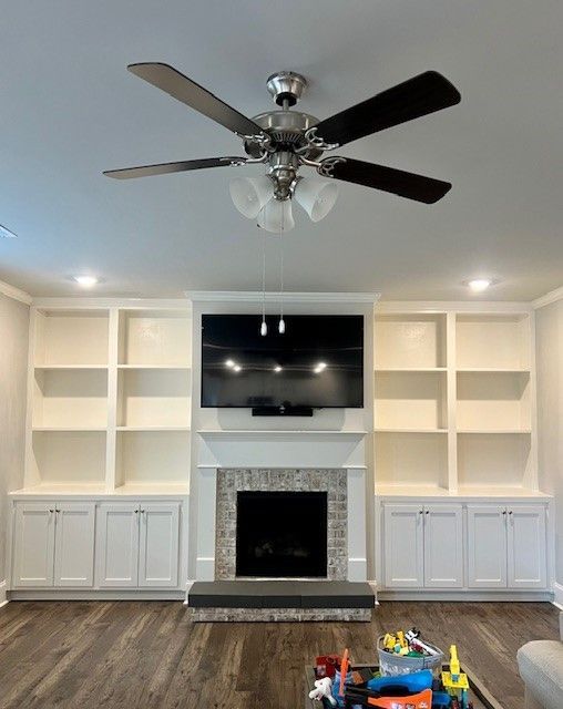 Living room with built-in shelves, fireplace, wall-mounted TV, and ceiling fan; hardwood floor and toys in foreground