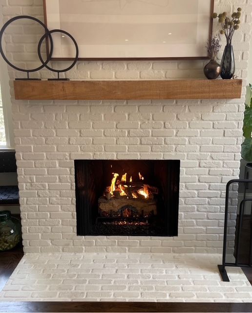 White brick fireplace with wood mantel, lit fire, and decorative metal rings and vase above.