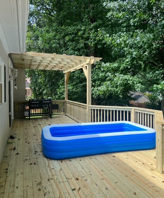 Wooden deck with pergola and blue inflatable pool beside a house, surrounded by trees.