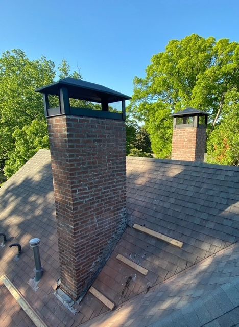 Rooftop with two brick chimney stacks under blue sky, surrounded by green trees.