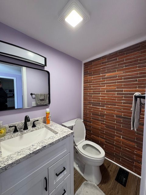 Small bathroom with lavender walls, brick accent wall, vanity sink, mirror, and toilet.