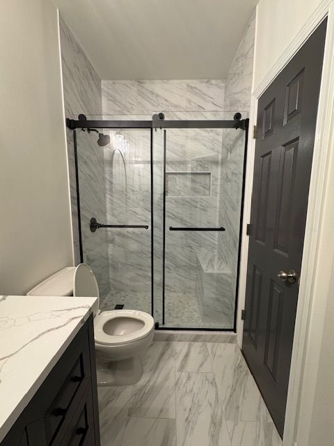 Narrow bathroom with marble tile, black vanity, toilet, and glass walk-in shower.