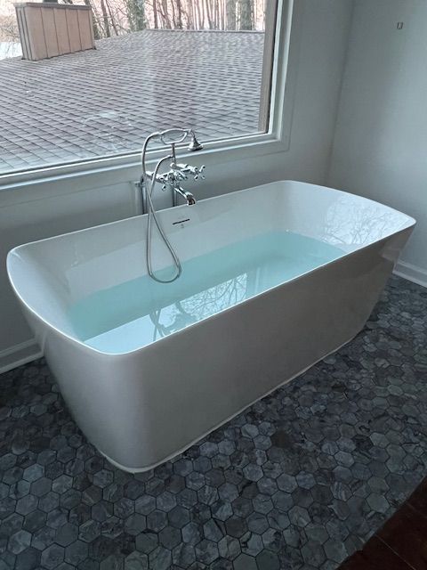 Modern white freestanding bathtub filled with water beside a large window.