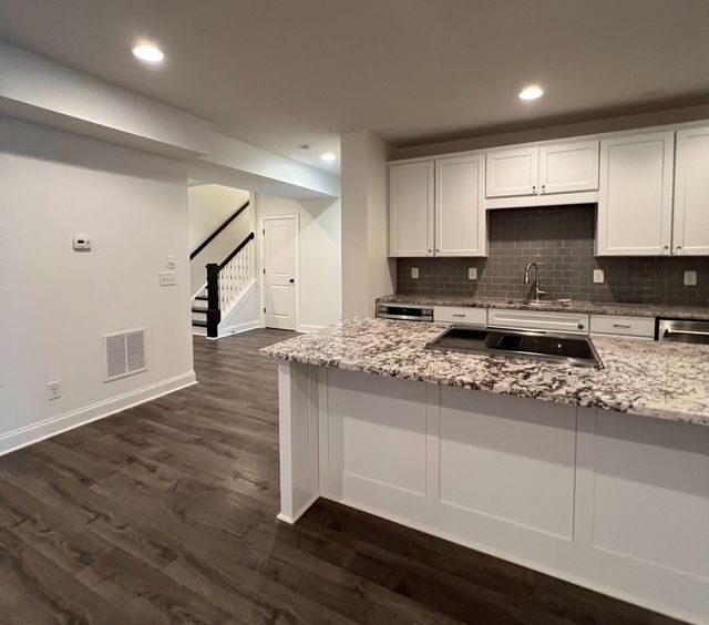 Modern open-plan kitchen with white cabinets, granite island, dark wood floors, and staircase in background