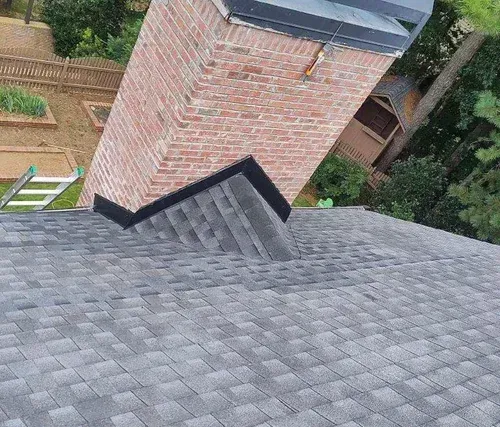 Gray shingled roof with a brick chimney and surrounding green trees and wooden fence