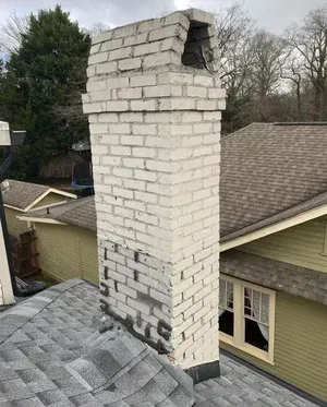White brick chimney on a shingled roof, with visible cracks and damage near the top.
