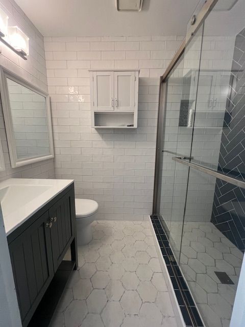 Modern bathroom with white tile, dark vanity, hex floor, toilet, and glass shower enclosure
