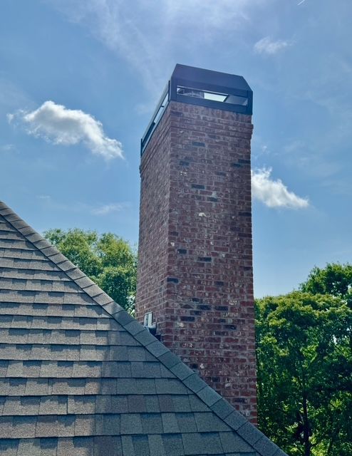 Brick chimney tower beside a shingled roof, with trees and blue sky in the background