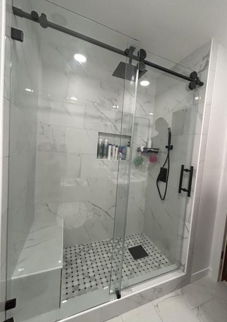 Glass walk-in shower with white tile floor, black hardware, and a built-in shelf with toiletries