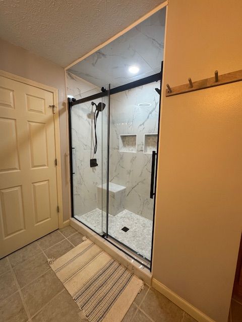 Bathroom with glass walk-in shower, beige tile floor, striped bath mat, and wall hooks.