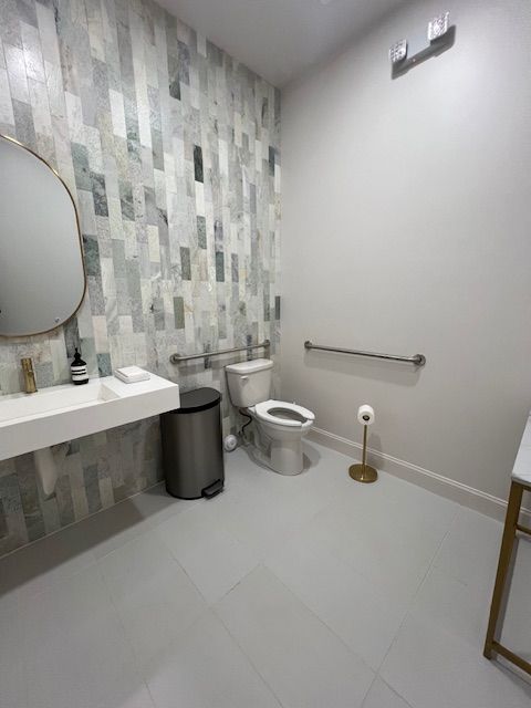 Small modern bathroom with sink, toilet, trash can, grab bar, and tiled wall.