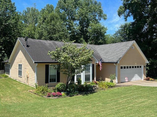 Single-story beige house with gray roof, attached garage, and U.S. flag in a sunny green yard.