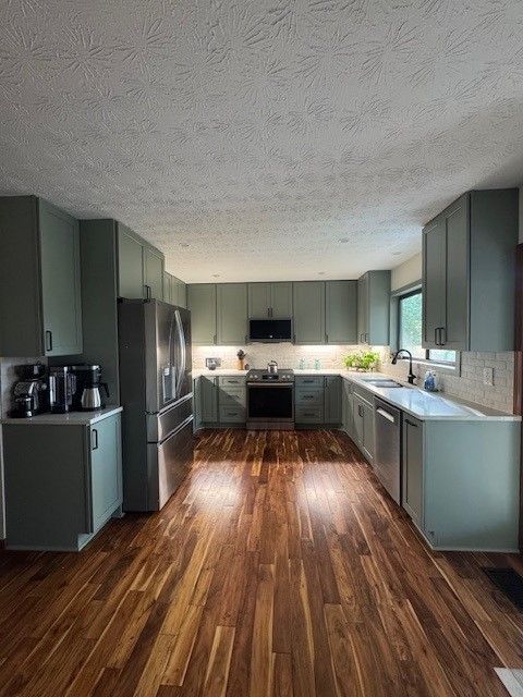 Wide kitchen with teal cabinets, stainless appliances, and wood-pattern floors in a bright room.