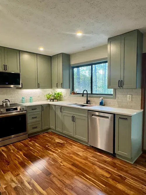 Modern kitchen with sage cabinets, stainless appliances, wood floor, and window above the sink