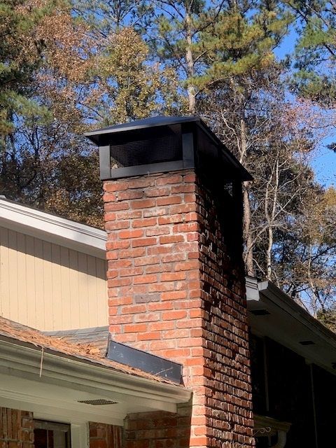 Brick chimney with black metal cap on a house roof, framed by autumn trees and blue sky