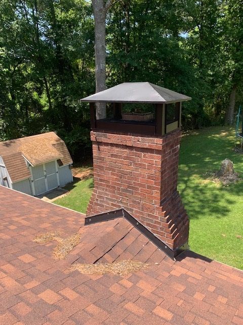 Brick chimney with metal cap on a shingled roof, overlooking a green backyard and trees.