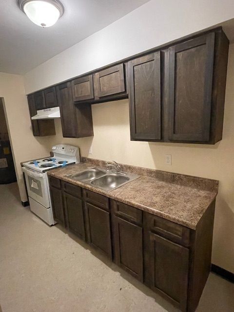 Small kitchen with dark wood cabinets, brown countertops, white stove, and double sink.