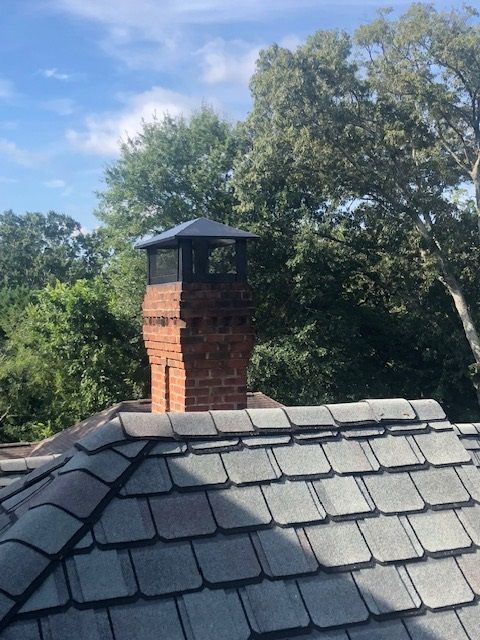 Slate roof with brick chimney and black cap against a blue sky and trees