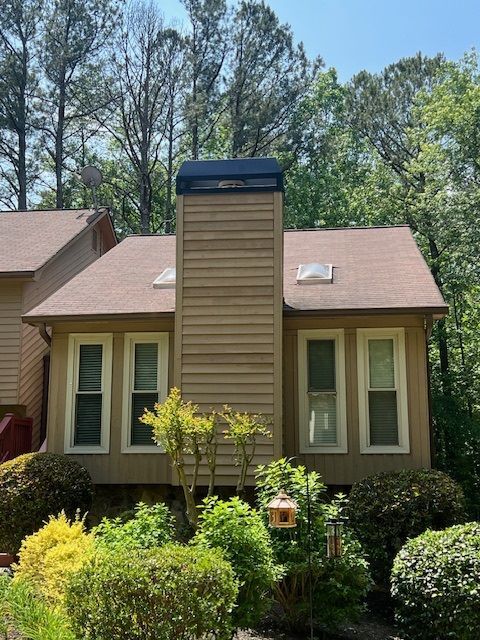 Suburban house with tan siding, central chimney, and a landscaped front yard under trees