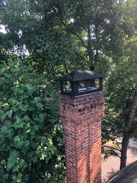 Brick chimney with a black cap rising above dense green trees