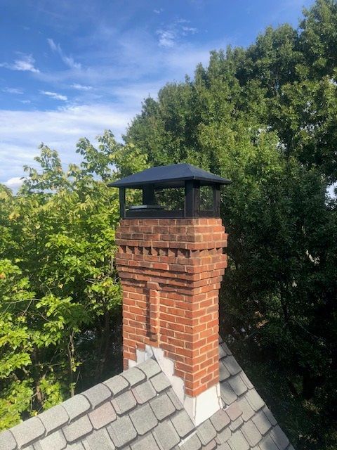 Brick chimney with black cap rising above a gray shingle roof against green trees and blue sky