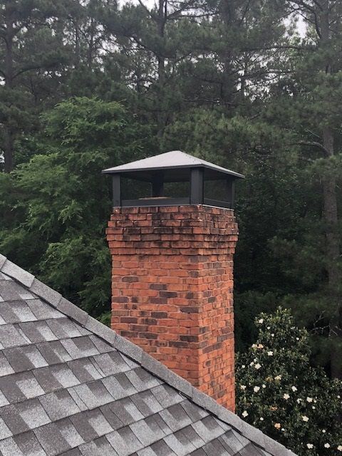 Brick chimney with black cap above a gray shingled roof, with trees in the background