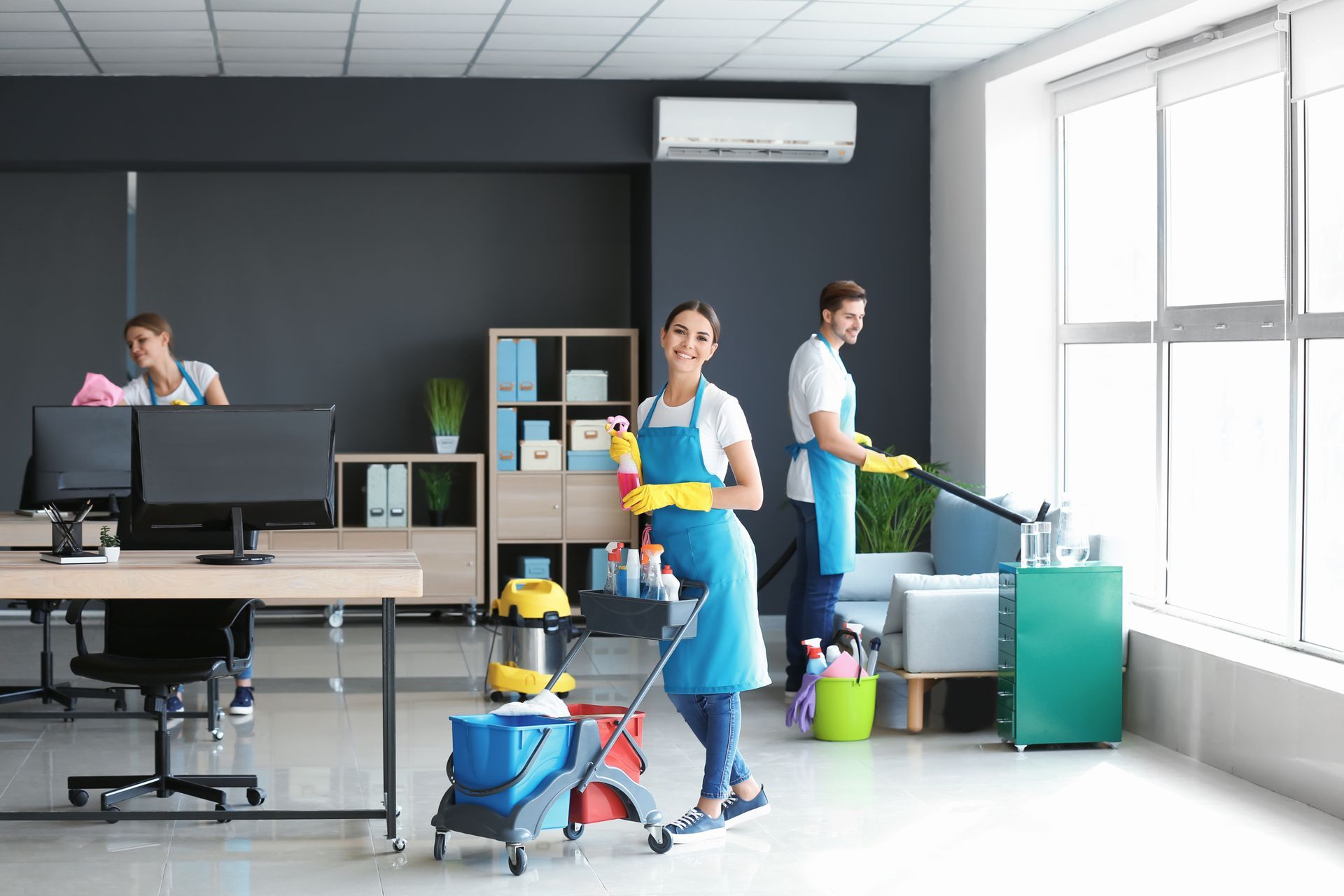 Office cleaning staff mopping a bright modern workspace with desks, carts, and cleaning supplies
