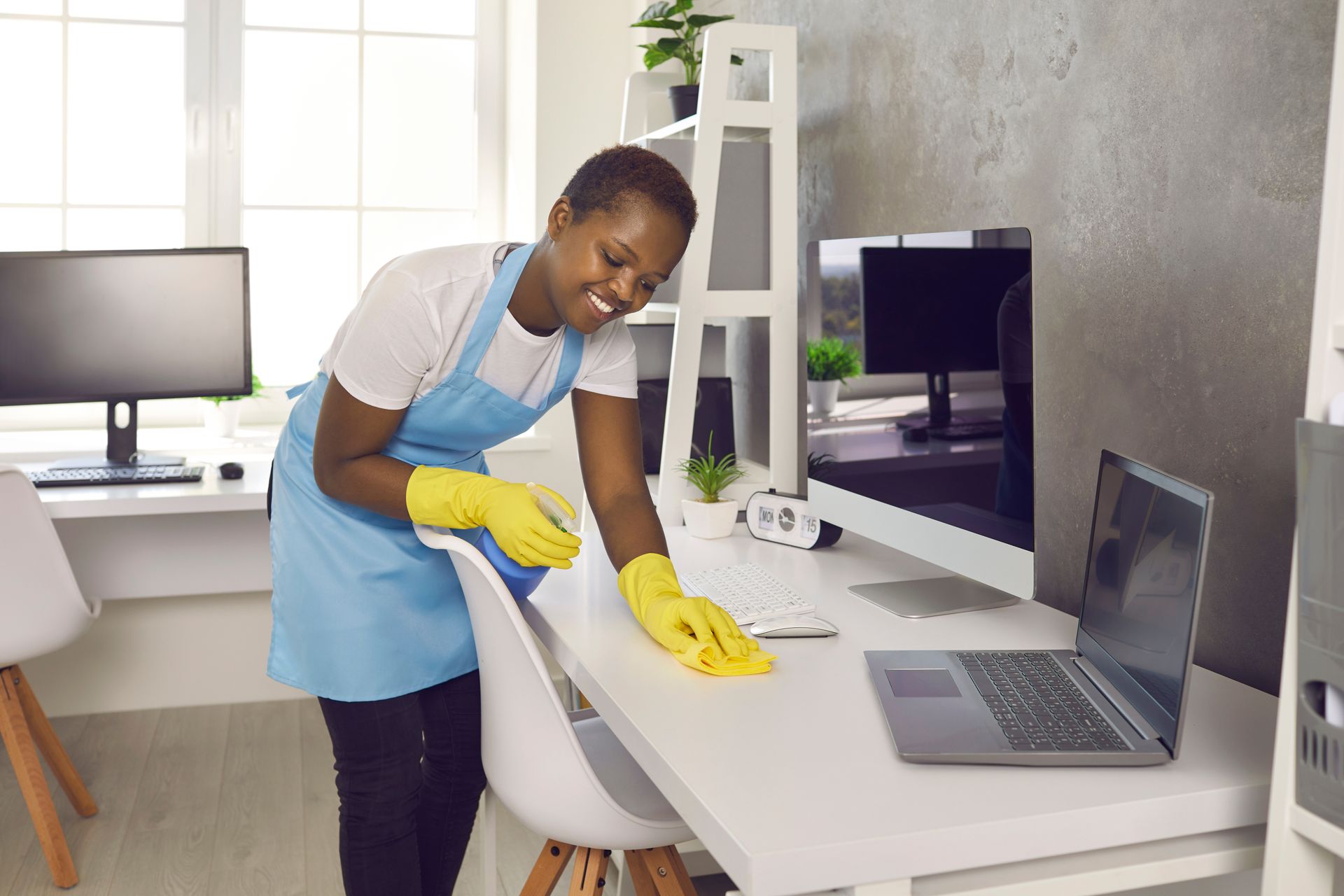 Person in blue apron cleaning a white desk with yellow gloves in a bright office.