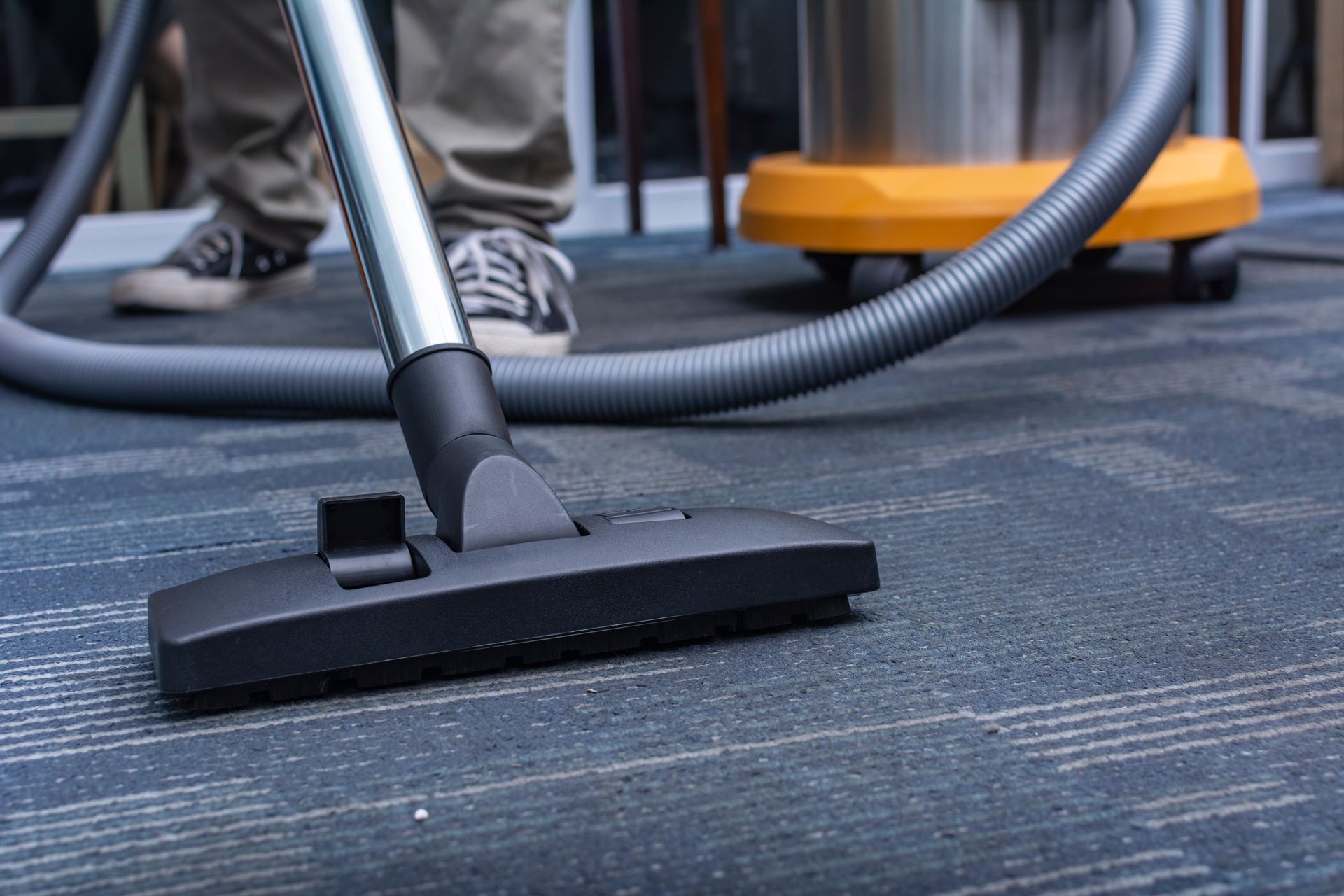 Vacuum cleaner head on a carpeted floor beside a person’s shoes and the vacuum canister Vacuum cleaner head on a carpeted floor beside a person’s shoes and the vacuum canister