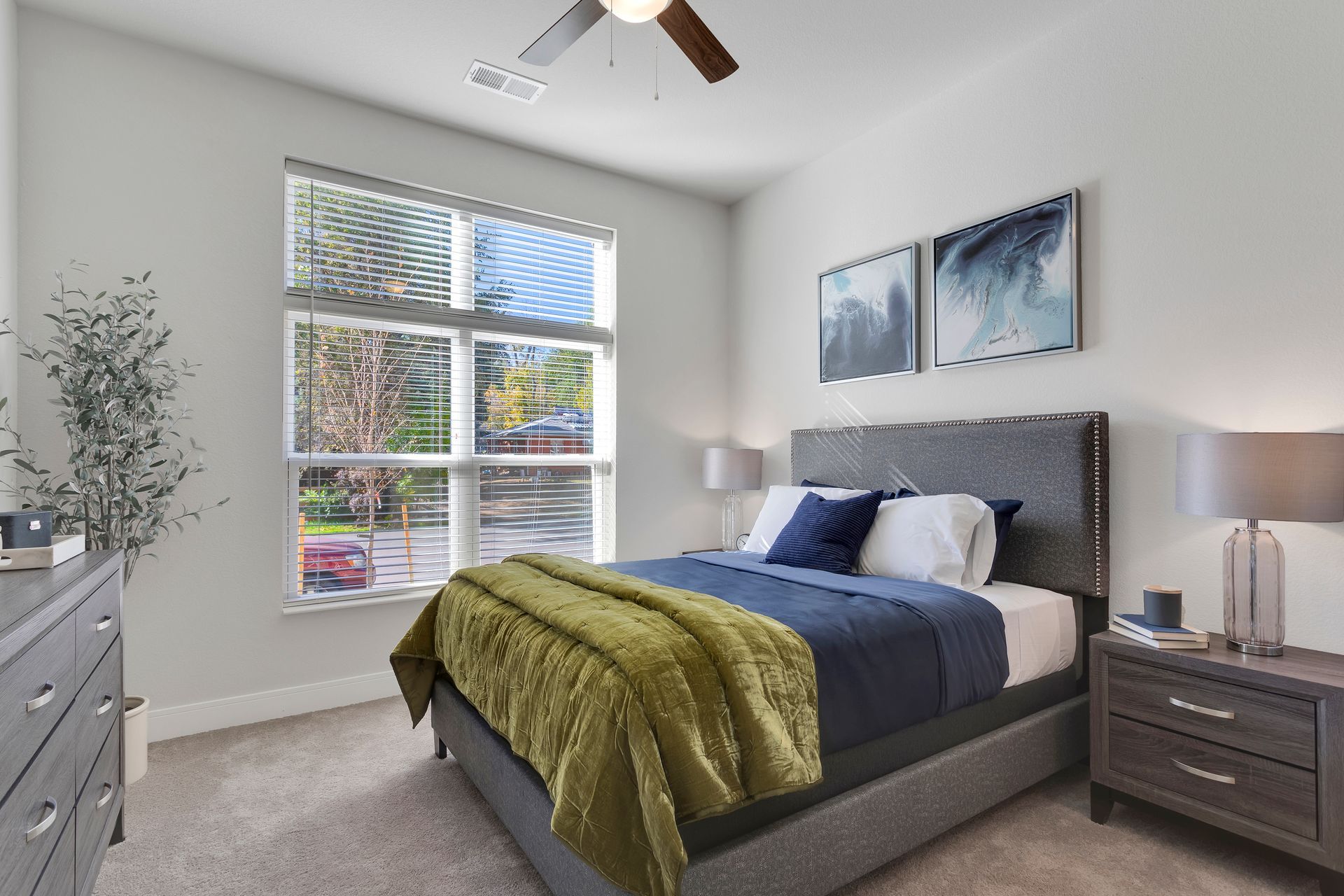 Bright bedroom with a gray bed, blue bedding, green blanket, large window, and ceiling fan.