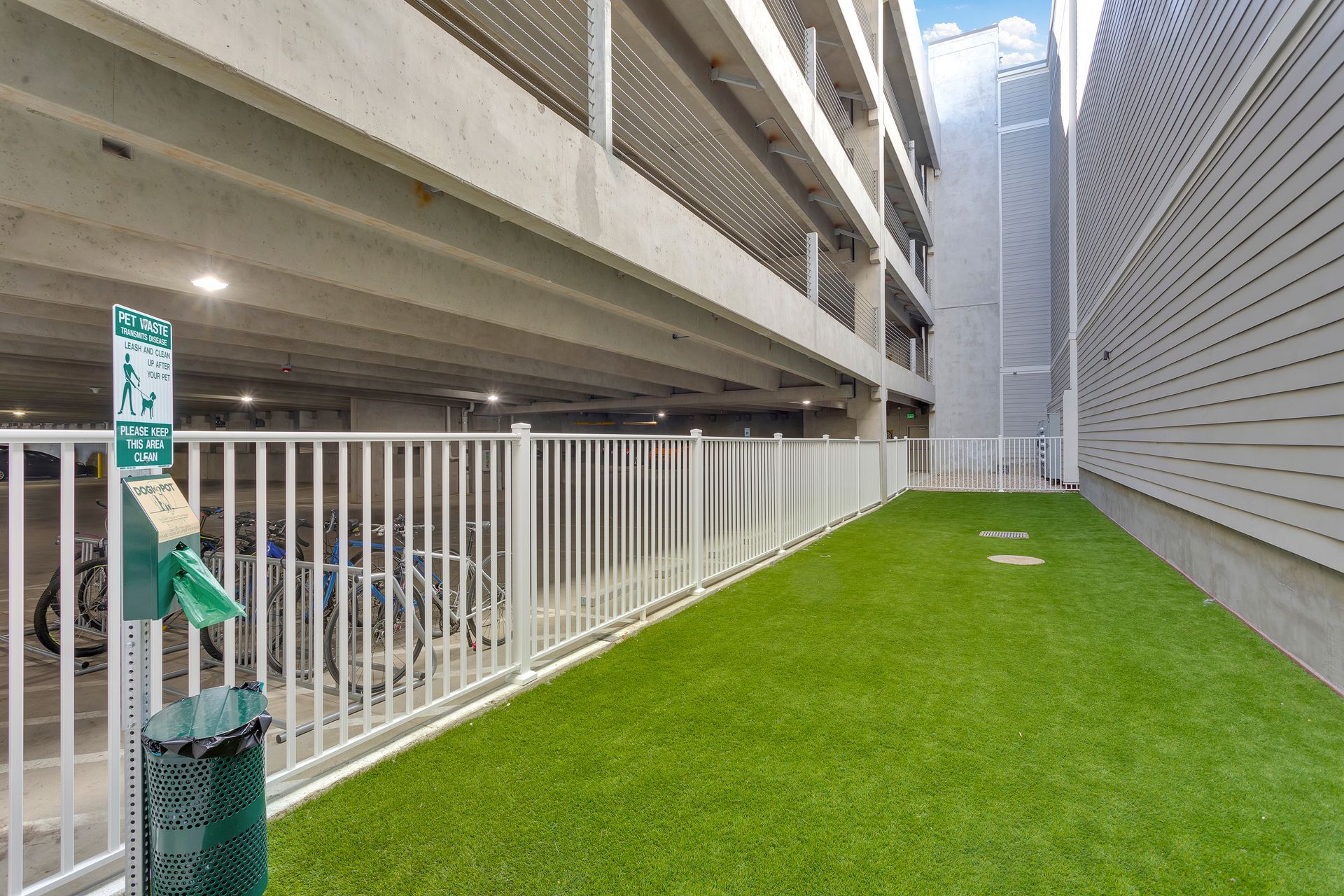 Narrow grassy corridor between tall buildings, with white railing and gray wall.
