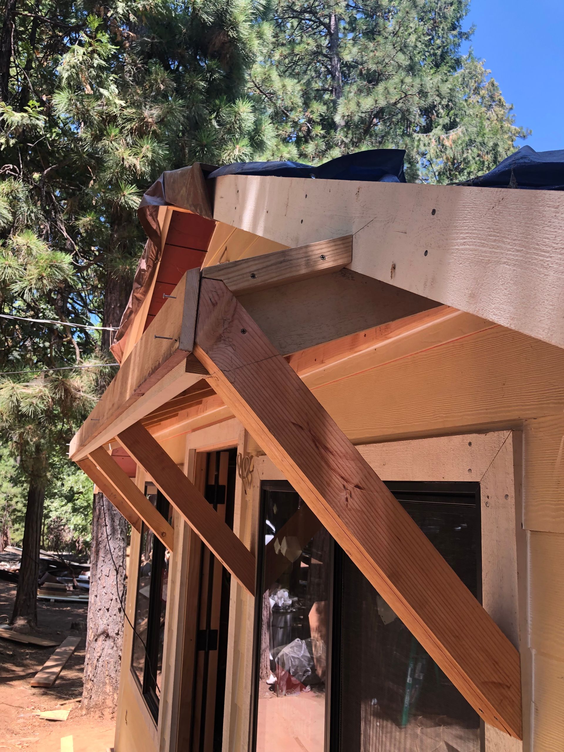 A wooden door awning under construction on a house exterior, with exposed framing and plywood set against a forest backdrop.
