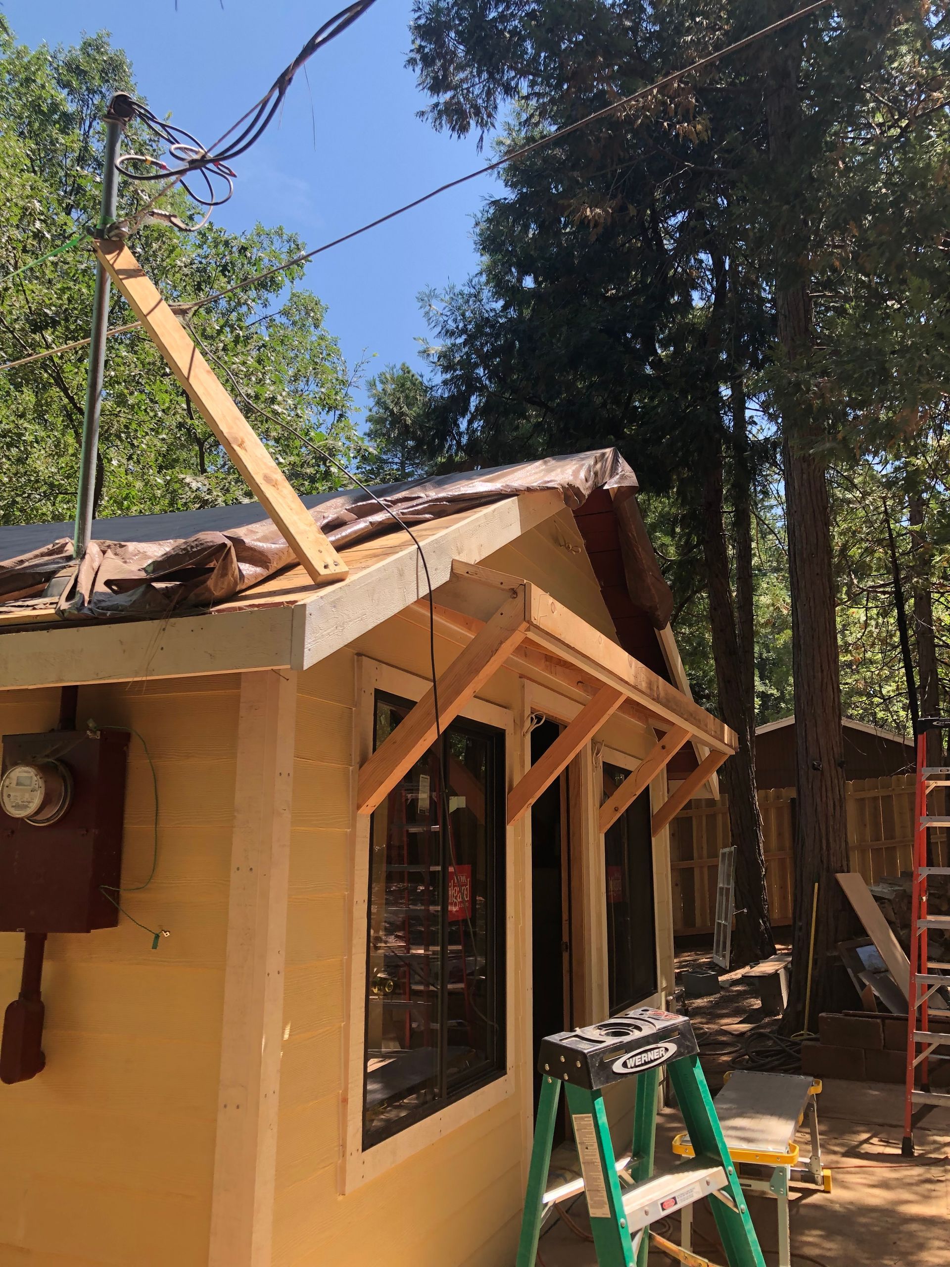 Yellow structure under construction with a temporary wooden roof support, electrical service mast, and a step ladder nearby.