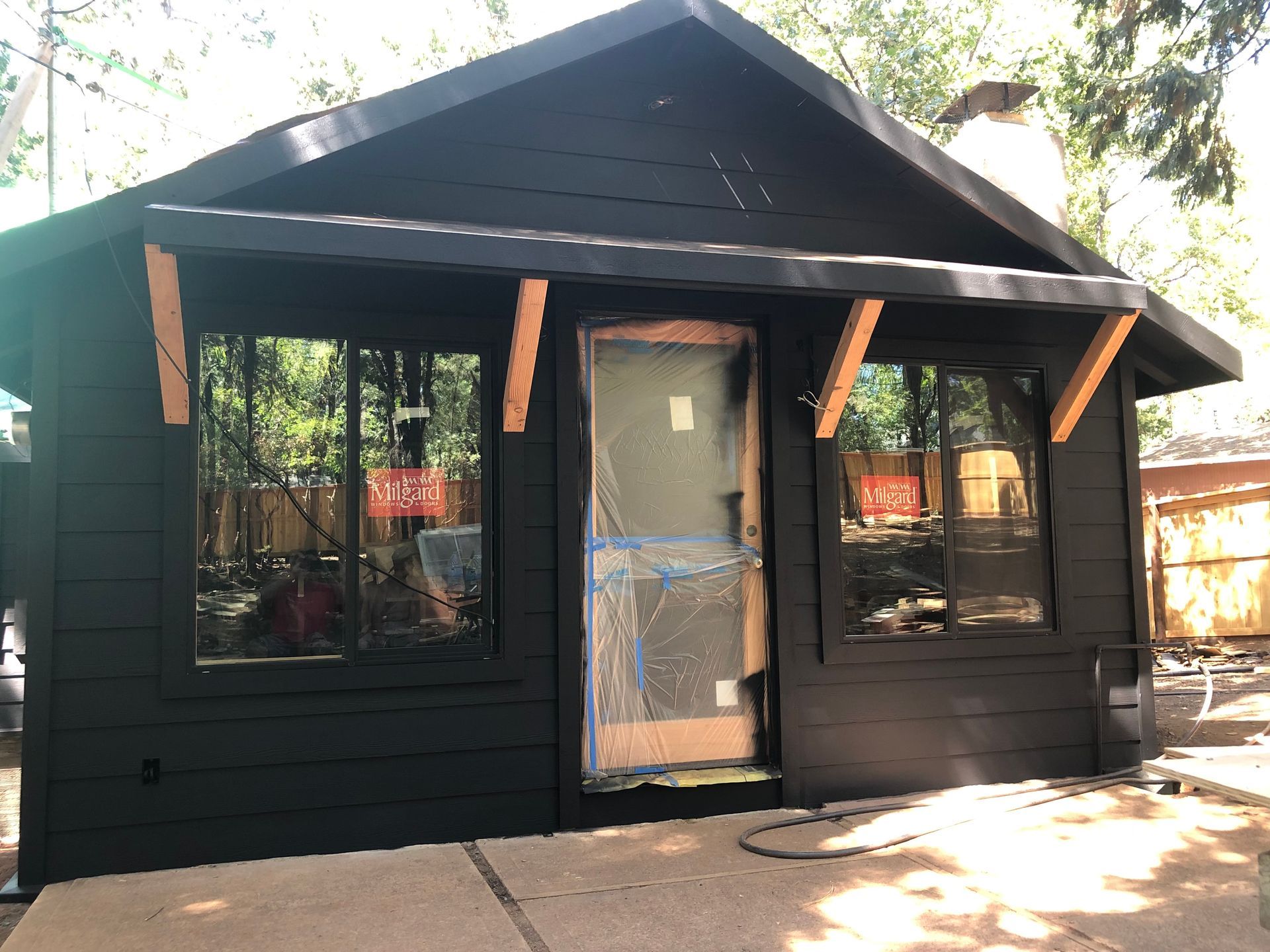 A small, dark-painted cabin with a front door, two windows, and wooden support beams for an awning, set in a yard.
