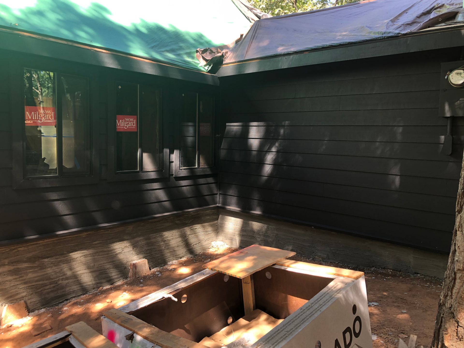 A construction site showing black exterior horizontal siding on a house with a framed, recessed foundation in the dirt yard.